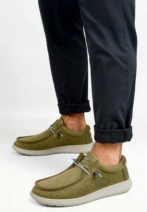 Chaussures en tissu vert avec une texture lisse, bout arrondi, lacets élastiques et une semelle gris clair. Pantalons noirs retroussés visibles.