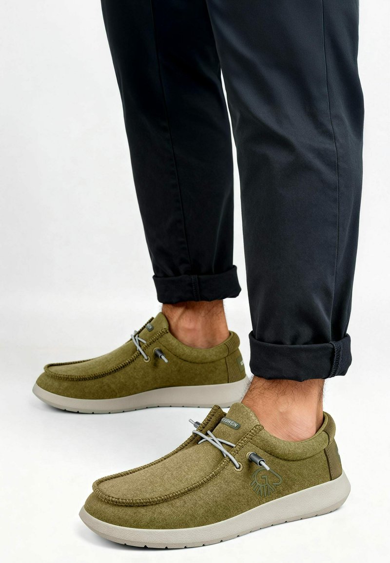 Chaussures en tissu vert avec une texture lisse, bout arrondi, lacets élastiques et une semelle gris clair. Pantalons noirs retroussés visibles.
