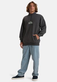 Sweatshirt zippé gris avec une poche, des poignets côtelés, et un logo "Billabong" vert sur la poitrine, associé à un pantalon bleu clair ample.