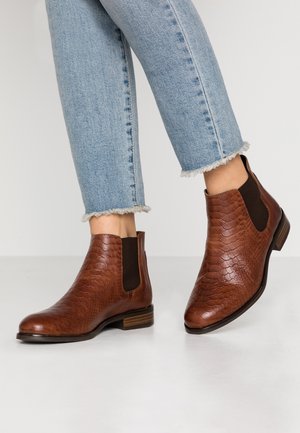 Botas Chelsea de cuero marrón con un patrón texturizado de cocodrilo, paneles laterales elásticos y un tacón de madera. Combinadas con unos pantalones vaqueros desgastados de corte corto en azul claro.