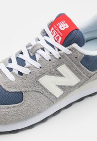 Zapatillas en gris y marino con materiales de ante y malla, que cuentan con cordones blancos, acentos de cuero y una etiqueta roja con "574" en la lengüeta.
