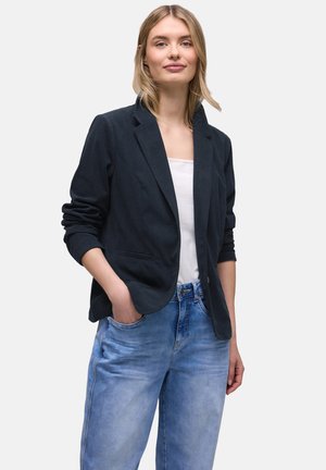 Blonde vrouw die een zwarte blazer draagt over een wit shirt en lichtblauwe spijkerbroek, staand met één hand in haar zak tegen een witte achtergrond.