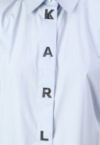 Camicia button-up azzurro chiaro con colletto strutturato. Le lettere argento "K," "A," "R," e "L" sono esposte lungo il piatto anteriore.