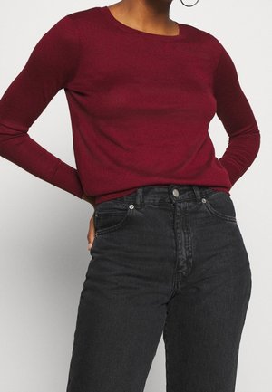 Personne à la peau foncée portant une chemise ajustée à manches longues bordeaux et un jean noir taille haute, debout les mains sur les hanches.