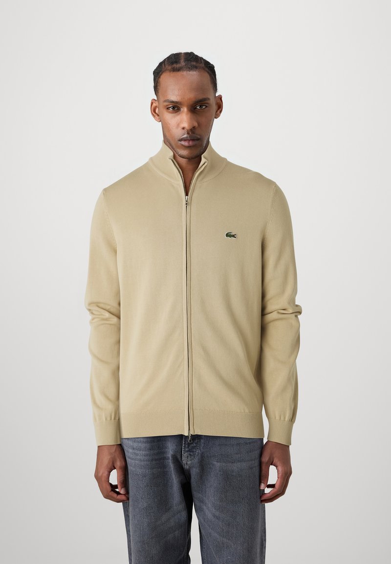 Šviesiai ruda zip-up cardigan megztas iš trikotažo, pasižymintis aukštu apykaklėje ir raukšlėtomis rankogaliais. Žalios spalvos logotipo akcentas kairėje krūtinės pusėje.