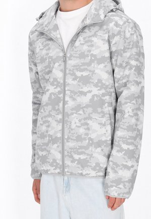 Mand iført lys grå-hvid digital camouflage lynlåstrøje med hætte, lyseblå jeans og hvid T-shirt.