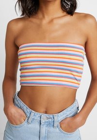 Persoon in een kleurige horizontaal gestreepte tube-top en lichtblauwe high-waisted jeans met handen in de zakken, torso zichtbaar.