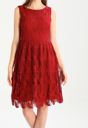 Robe rouge en dentelle sans manches avec un corsage ajusté et une jupe évasée, portée par une personne debout sur un fond uni.