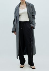 Grijze pinstriped lange jas over een witte tanktop, gecombineerd met zwarte cargobroeken met een zak en zwarte open-toe haksandalen.