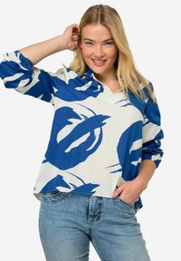 Blusa blu e bianca con motivo floreale, dotata di scollatura a V, maniche lunghe a sbuffo e vestibilità ampia, abbinata a jeans in denim azzurro chiaro.