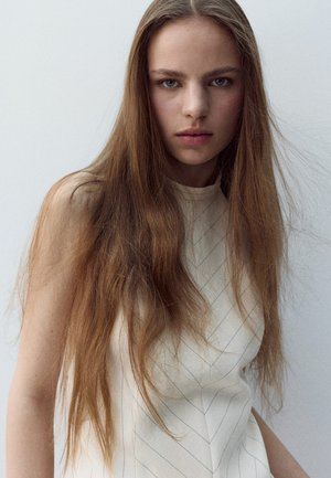 Young woman with long brown hair wearing a sleeveless white top, looking directly at the camera against a plain light background.