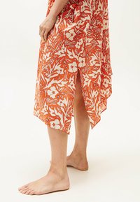 Vestito caftano floreale arancione con orlo asimmetrico, realizzato in tessuto leggero, caratterizzato da motivi botanici bianchi e spacchi laterali.