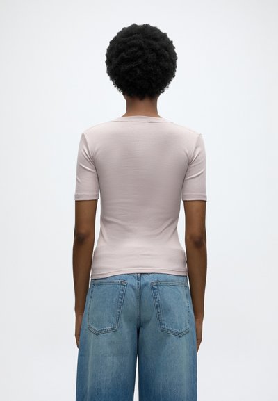 Άτομο με κοντά σγουρά μαλλιά που φορά ανοιχτό ροζ κοντομάνικη μπλούζα και ψηλόμεσο μπλε τζιν, εμφανίζεται από την πίσω πλευρά.