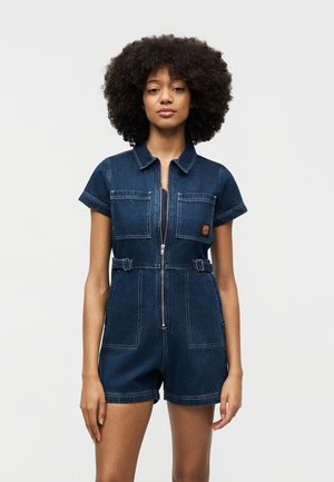 Femme aux cheveux bouclés portant une combinaison courte en denim foncé à manches courtes avec fermeture éclair à l'avant et poches poitrine, debout devant un fond uni.