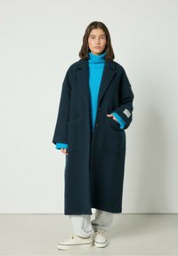 Námornícky vlnený kabát s nadmerným strihom, veľkými prednými vreckami a širokými klopami, preložený cez svetlomodrý rolák, v kombinácii so šedými teplákmi.