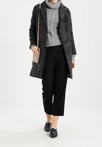 Abrigo largo con un patrón en negro y gris, suéter de punto en gris, pantalones recortados negros y zapatos negros sin cordones, junto con un bolso cruzado beige.