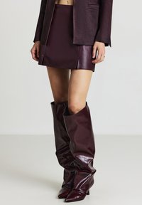 Mini gonna in ecopelle bordeaux abbinata a stivali alti morbidi fino al ginocchio, con punte affusolate e una finitura lucida e elegante.