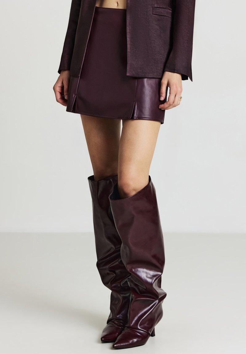 Mini gonna in ecopelle bordeaux abbinata a stivali alti morbidi fino al ginocchio, con punte affusolate e una finitura lucida e elegante.