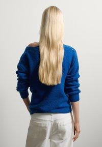 Suéter azul de punto con un escote amplio, tejido texturizado y puños acanalados, combinado con pantalones blancos. Cabello largo y liso visible.