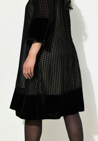 Schwarzes Samtkleid mit texturiertem Hahnentrittmuster, ausgestellten Ärmeln und einem einfarbigen dunklen Saum. Glattes, weiches Material mit glänzendem Finish.