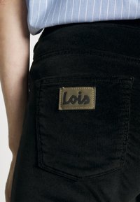 Black corduroy pants with a rear pocket featuring a rectangular leather patch labeled "Lois" in a cursive font. Striped shirt is partially visible.