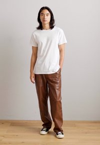 T-shirt blanc à manches courtes associé à un pantalon en cuir marron. Le pantalon a une texture légèrement brillante et une coupe décontractée. Des baskets noires complètent le look.