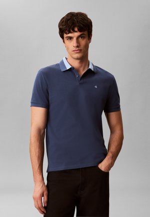 Młody mężczyzna z ciemnymi włosami, ubrany w granatową koszulkę polo i czarne spodnie, stoi z jedną ręką w kieszeni na jednolitym szarym tle.