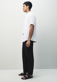 Weißes kurzärmeliges Leinenhemd, lockere schwarze Nadelstreifenhose und schwarze Sandalen mit doppeltem Schnallendetail, seitlich betrachtet.
