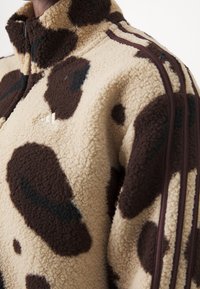 Fleece zip-up jacka med en tan bas, bruna och svarta oregelbundna fläckar, hög krage och bruna sidostraper. Har en adidas-logotyp.