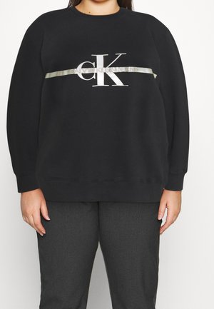 Czarna bluza z długim rękawem z dużym białym logo "cK" i srebrnym napisem "Calvin Klein Jeans" na piersi, noszona z ciemnoszarymi spodniami.