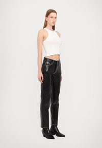 Calças de couro pretas com detalhe em costura branca, combinadas com um top cropped branco ajustado. A modelo está em botins de cano curto pretos com salto.