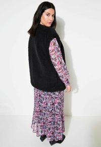 Schwarze, plüschige, ärmellose Weste über einem langen, blumigen Kleid mit rosa und blauen Mustern, wobei schwarze Stiefel am Boden sichtbar sind.