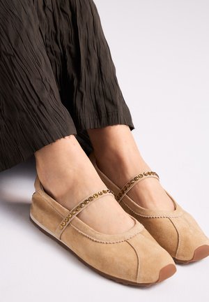 Zapatos planos Mary Jane de ante beige con una correa adornada con gemas, usados con pantalones sueltos oscuros y texturizados sobre un fondo blanco.