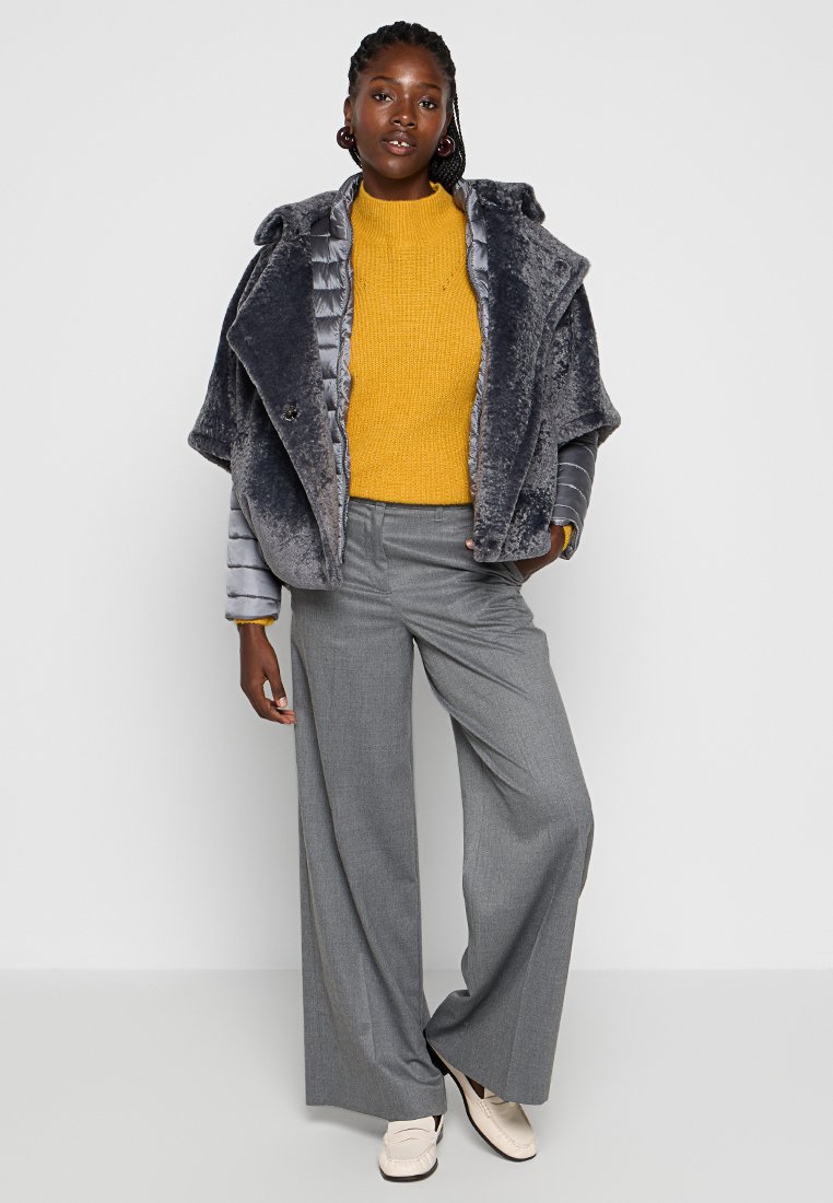 Grauer Kunstfellmantel mit einer gepolsterten Innenschicht, kombiniert mit einem leuchtend gelben Strickpullover, grauen weitem Hosen und beigen Schuhen.