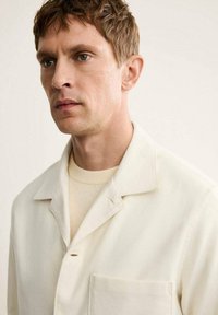 Camisa blanca de cuello en tejido suave, con un bolsillo con parche y un cierre de un solo botón. Llevada sobre un suéter de cuello redondo de color beige claro.