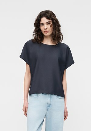 Femeie cu păr creț și întunecat, purtând un top larg cu mâneci scurte, de culoare bleumarin, și blugi cu talie înaltă deschis la culoare, stând în picioare pe un fundal simplu.