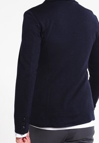 Navy blazer with a fitted design, textured fabric, and three-button accents on the sleeve. Smooth finish and tailored shape.