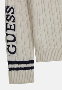 Maglione lavorato a maglia color avorio con motivo a trecce, polsini a coste e logo blu navy lungo la manica con dettagli a righe a contrasto.