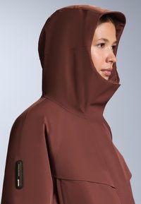 Barna vízálló dzseki magas gallérral és kapucnival. Texturált anyagból készült, és a ujján egy cipzáras zseb található tárolásra.