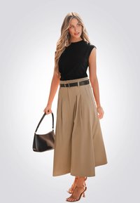 Top negro de manga corta, pantalones beige de tiro alto con pliegues, cinturón negro, sosteniendo un bolso negro y usando sandalias de tacón color marrón.
