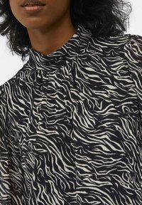 Blouse noire avec imprimé zèbre blanc, dotée d'un col haut et de détails froncés au niveau du cou. Tissu léger avec une texture lisse.
