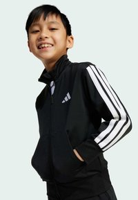 Černá zipová bunda s bílými pruhy na rukávech, s vysokým límcem, předními kapsami a logem adidas na hrudi.