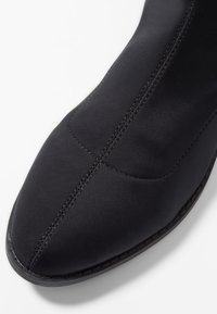 Bota de tela negra con una textura suave, puntera afilada y costuras visibles a lo largo de las costuras, con una suela duradera para mayor estabilidad.