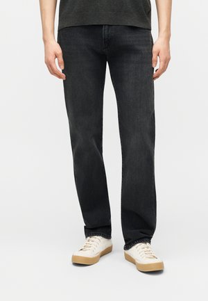 Osoba ubrana w proste czarne dżinsy i białe trampki z gumowymi podeszwami, stojąca na tle jednolitego jasnego tła.