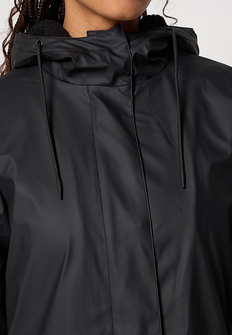 Veste noire imperméable avec capuche, cordons de serrage réglables et fermeture éclair frontale dissimulée, portée par une personne avec des cheveux tressés.