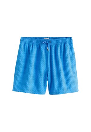 Primo piano di una persona che indossa pantaloncini blu con motivo, con tasca posteriore e un braccialetto di perline al polso destro.