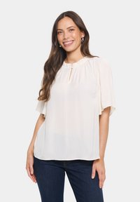 Krämfärgad blus med volangkrage och korta, svävande ärmar. Har en nyckelhålsdetalj fram och är tillverkad av lätt tyg.