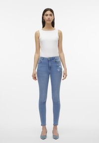 Blugi skinny din denim albastru deschis cu detalii brodate florale pe coapsă, purtați cu un top alb ajustat și tocuri albastre deschise.