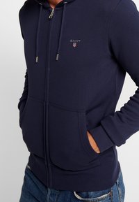Marinblå zip-up hoodie i mjuk bomull, med en känguruficka, ribbade ärmslut och en liten broderad logotyp på bröstet.