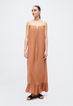 Femme aux cheveux plaqués en arrière portant une robe maxi sans bretelles, ample, de couleur rouille avec un ourlet à volants et des sandales à enfiler noires, debout sur un fond uni.